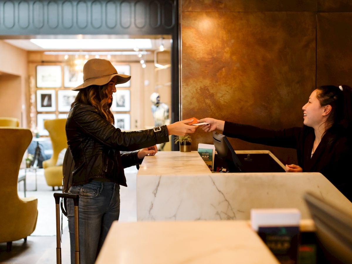 A woman is checking into a hotel, handing a card to the receptionist at a marble counter, with luggage beside her in a well-lit lobby.
