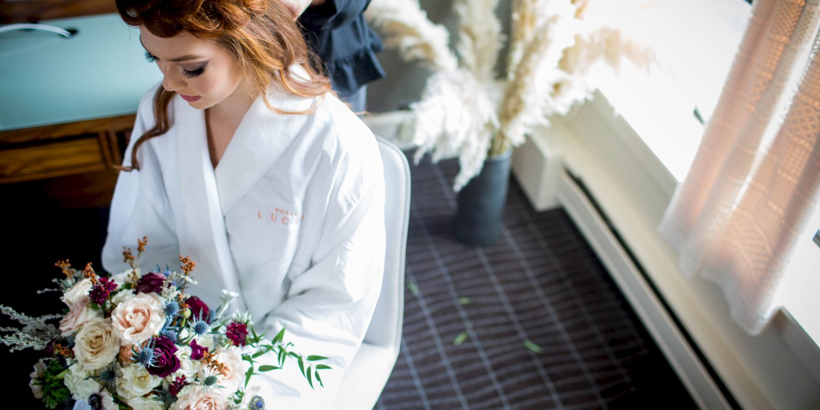 A woman in a white robe holds a bouquet while her hair is styled, with flowers and a window in the background.