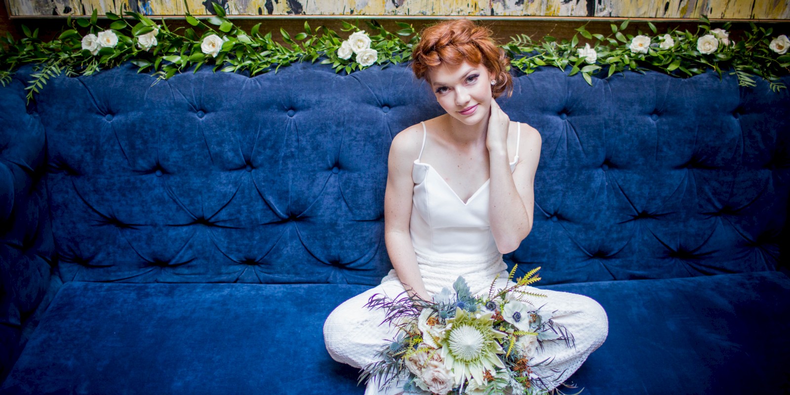 A person with red hair sits on a blue couch holding a bouquet of flowers, with green leaves and white flowers in the background.