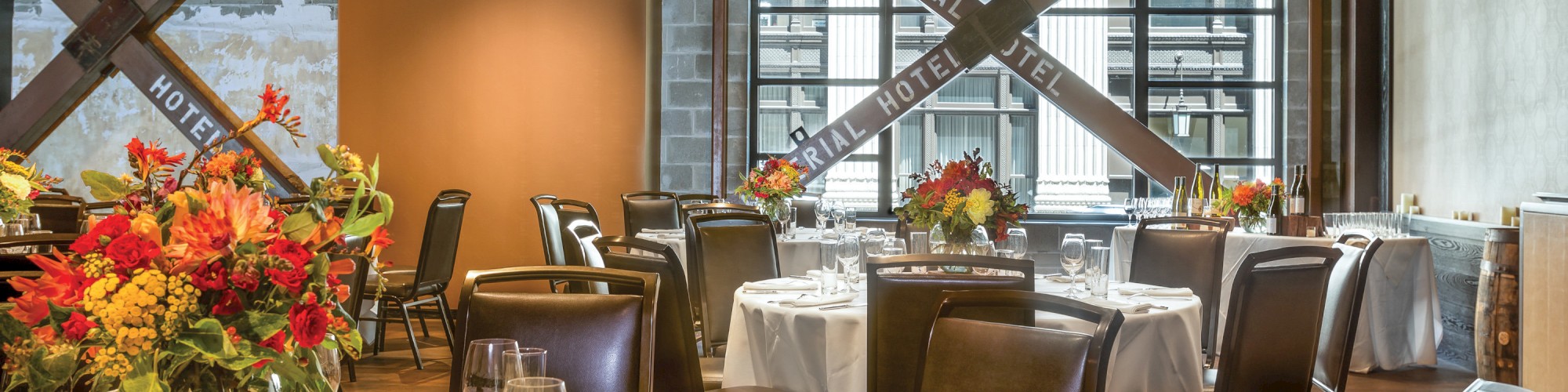 A beautifully arranged dining area with round tables, floral centerpieces, and wooden chairs, set in a spacious, elegant room with large windows.