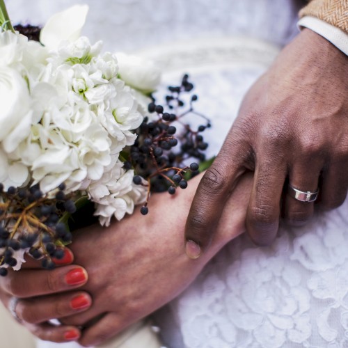 A close-up of a couple holding hands, featuring wedding rings and a bouquet of white flowers with dark berries, taken on their wedding day.