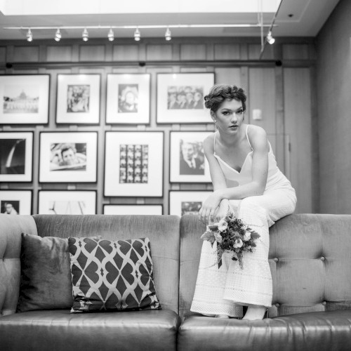 A person in a white dress sits on a couch holding a flower bouquet in a room with framed pictures on the wall.