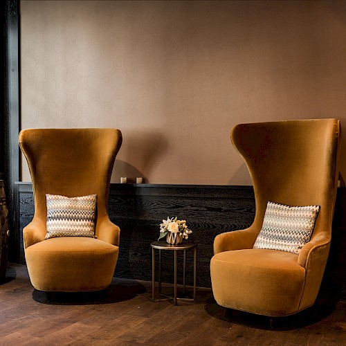 A stylish hotel lobby with large windows, velvet armchairs, a barrel table with flowers, and a decorative beam displaying 