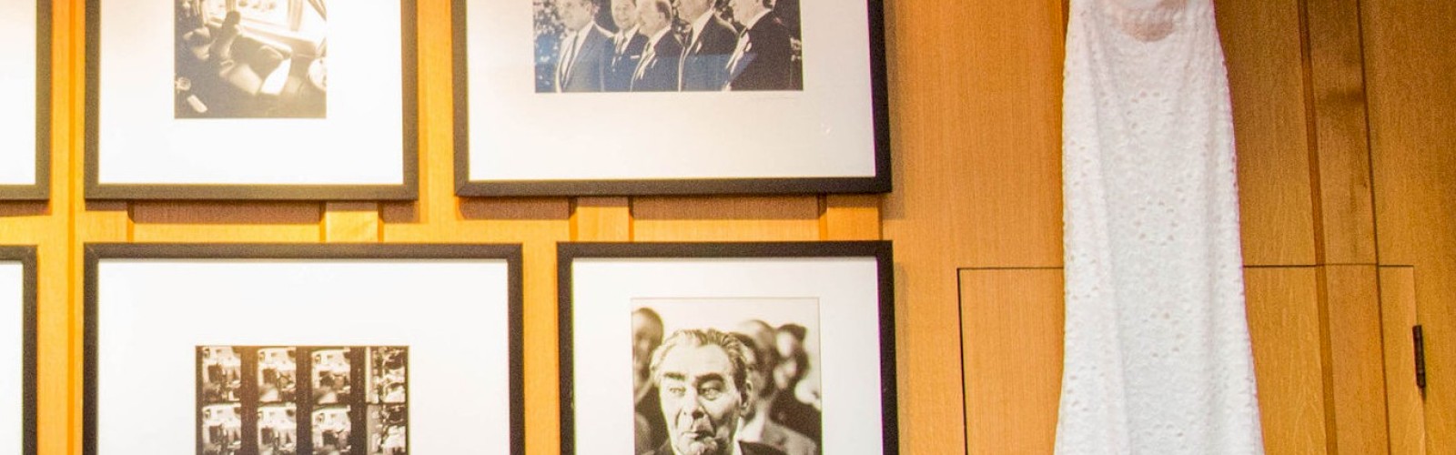 A white dress hangs on a wooden wall next to black-and-white framed pictures. There are flowers at the bottom right corner of the image.