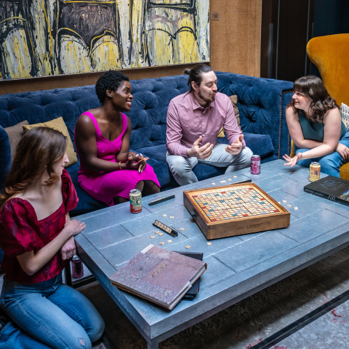 Four people are sitting around a table with a Scrabble game, enjoying drinks and a lively conversation in a cozy room setting.