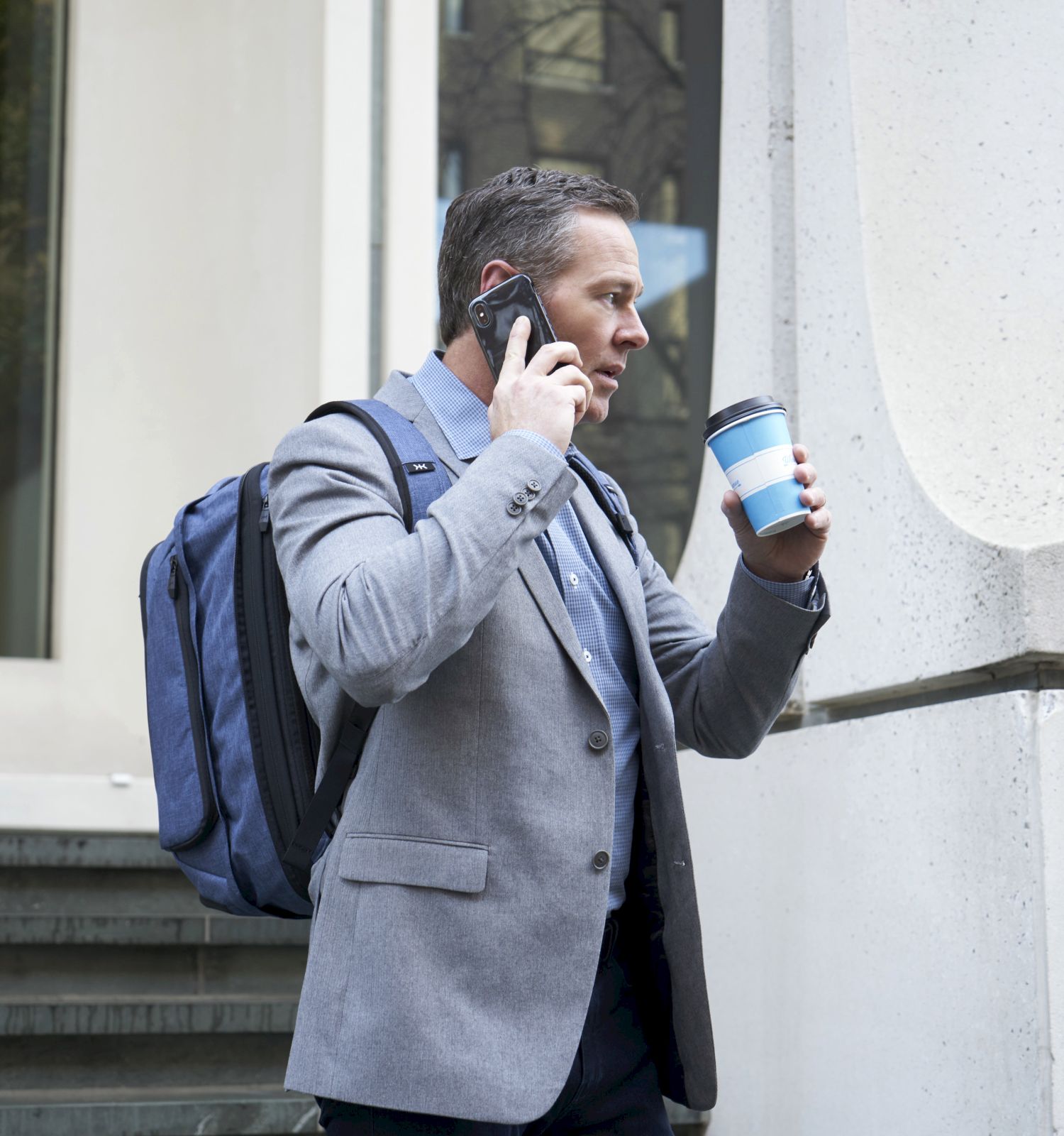 A man in a gray blazer with a backpack talks on the phone and holds a coffee cup, standing near a building with glass windows and steps.
