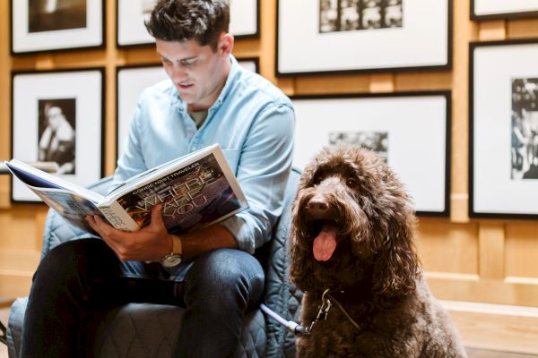 A man wearing a light blue shirt reads a book while a brown furry dog sits beside him; black-and-white photos hang on the wall behind them.