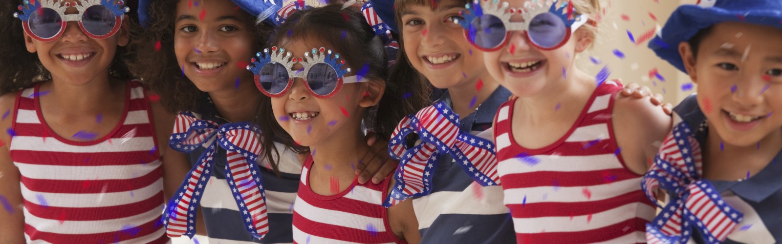 A group of children wearing red, white, and blue outfits, along with patriotic hats and glasses, are smiling and standing together indoors.