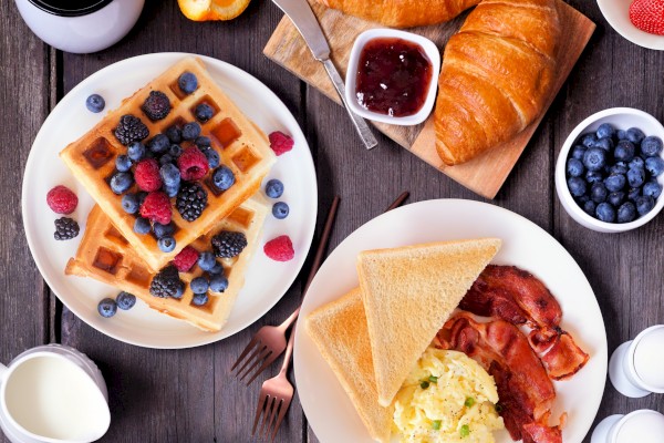 A breakfast spread with waffles, berries, croissants, jam, toast, scrambled eggs, bacon, blueberries, orange slices, milk, and coffee.