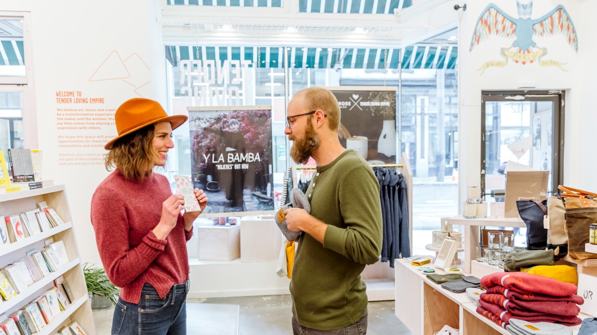 Two people are talking inside a store with shelves of books and clothes around them. There's natural light coming in through large windows.