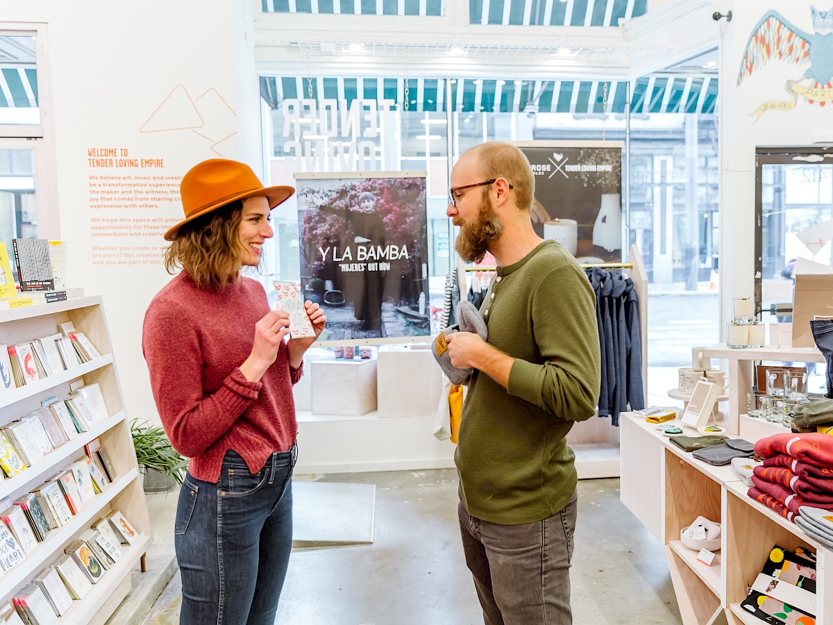 Two people are talking inside a store with shelves of books and clothes around them. There's natural light coming in through large windows.