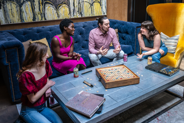 Four people are sitting around a table playing a board game, with a colorful painting in the background.