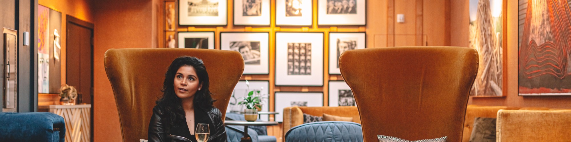 A person sits in a stylish, modern lounge with framed art, holding a glass, surrounded by cozy furniture and warm lighting.