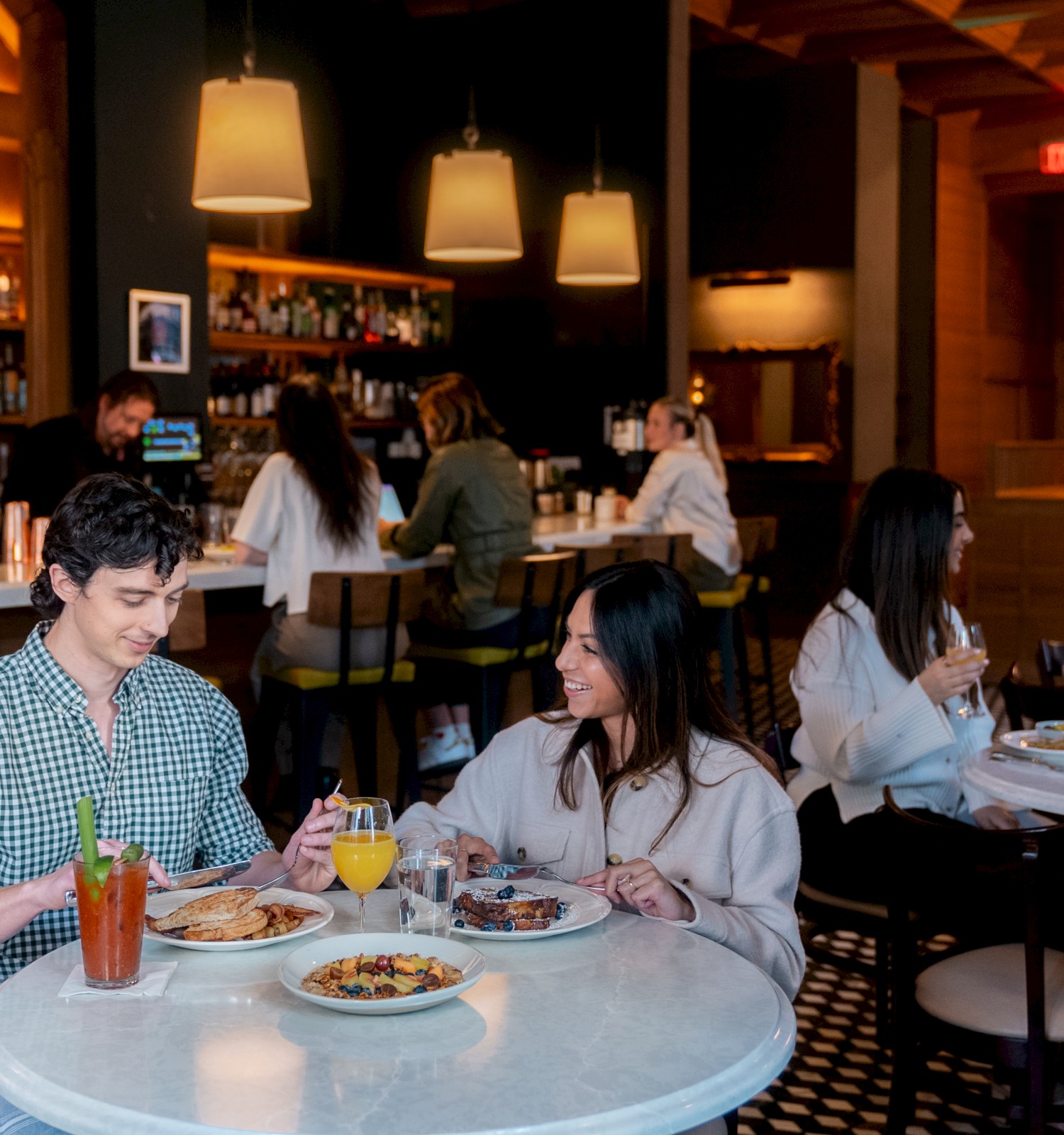 People dining in a cozy restaurant with a bar, enjoying their meals and drinks at round tables.