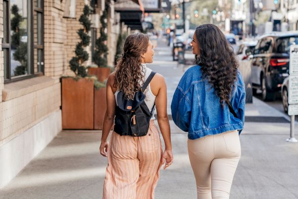 Two women are walking down a city street together, one wearing a backpack and the other a denim jacket. They are approaching a building with an overhang.