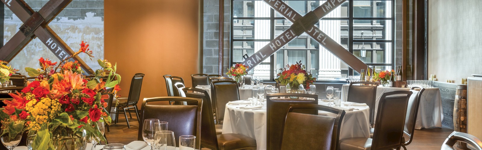 The image shows a decorated dining area with round tables, chairs, floral centerpieces, and large windows displaying the words 