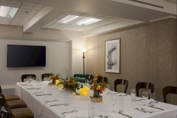 A conference room with a long table set for dining, featuring floral centerpieces, candles, a wall-mounted TV, and framed artwork.