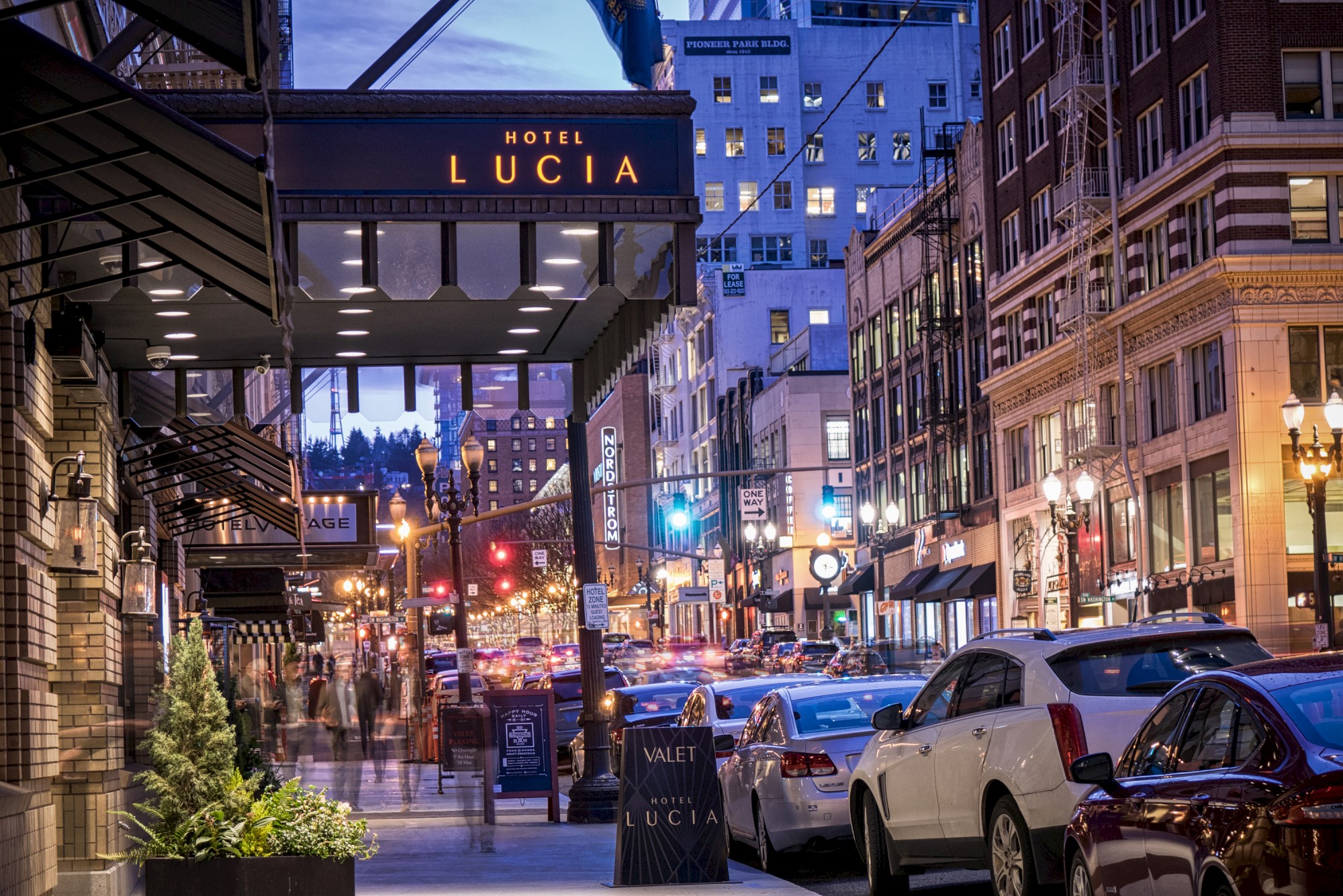 Busy urban street scene at dusk with cars, pedestrians, and Hotel Lucia's sign illuminated. Buildings line the street amid city lights.