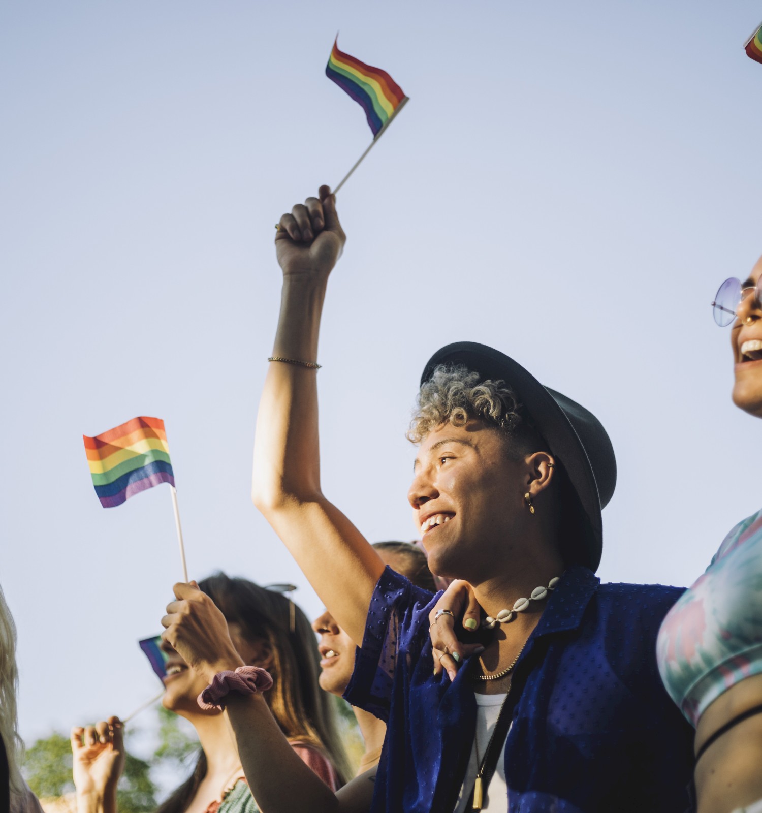 A group of people is celebrating outdoors, joyfully waving rainbow flags while smiling and cheering in support of LGBTQ+ pride.