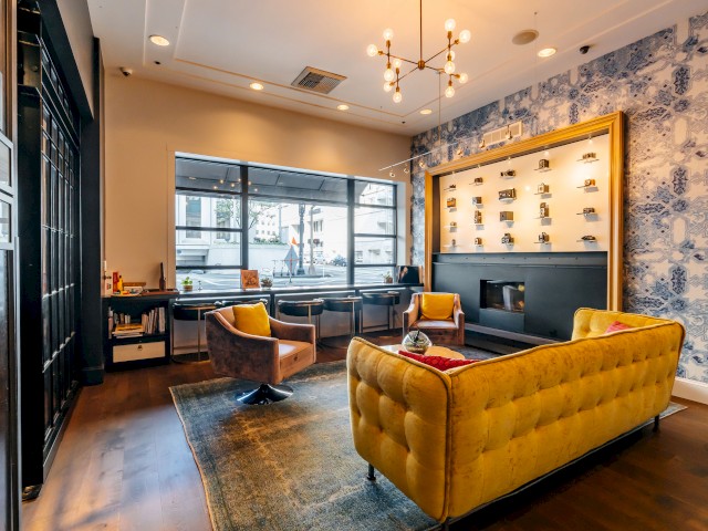 A stylish room with yellow seating, patterned wallpaper, large windows, a modern chandelier, and shelving displaying items.