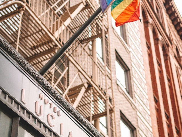 The image shows the exterior of Hotel Lucia with a rainbow flag attached to a pole, located in front of a building with fire escapes.