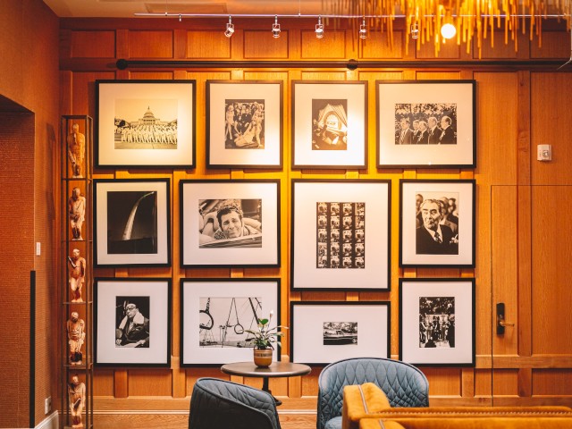 A well-decorated room with a wooden wall displays black-and-white framed photographs in a grid pattern, with blue armchairs in the foreground.