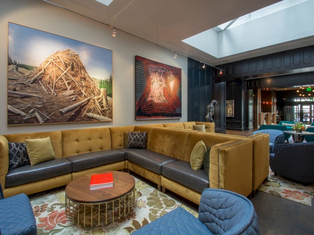 This image shows a modern lounge area with a sectional sofa, blue chairs, and a round wooden coffee table. Artworks are displayed on the wall.