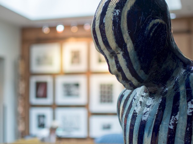 A striped sculpture of a human figure is in the foreground, with framed artworks displayed on a wall in the blurred background.