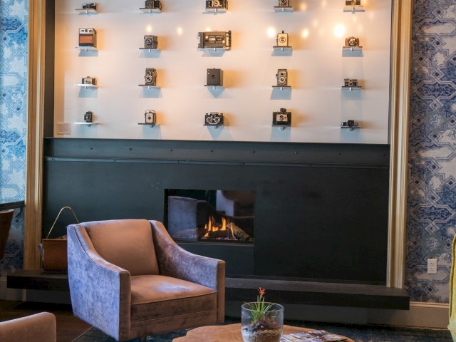 The image shows a cozy seating area with two armchairs, a wooden coffee table, and a fireplace. The wall has shelves displaying various objects.