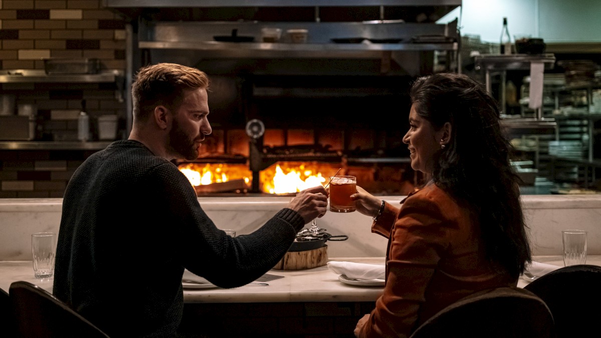 A couple sits at a restaurant counter, toasting with drinks in front of a lit wood-fired oven in a cozy setting.