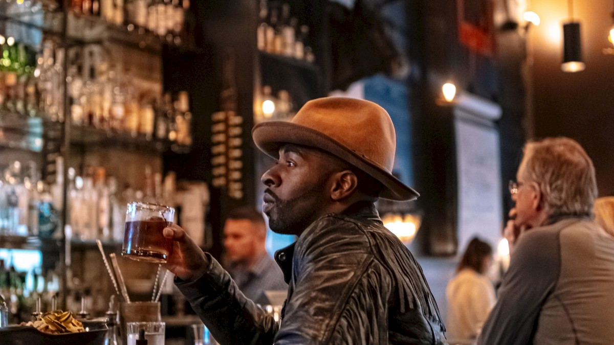 A man in a hat sits at a bar, holding a drink, with shelves of bottles in the background and dim lighting setting a cozy ambiance.