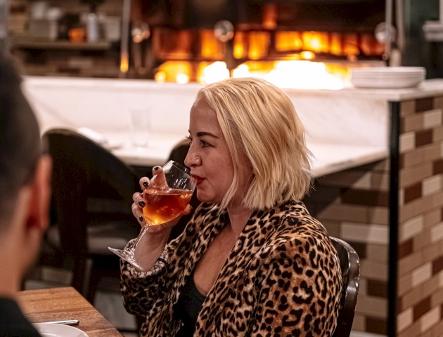 A person with blonde hair sips a drink in a restaurant with a glowing wood-fired oven in the background, wearing a leopard print jacket.