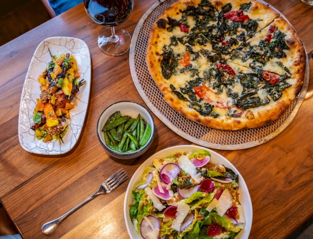 The image shows a table with a pizza, a dish with roasted vegetables, a bowl of snap peas, a salad with radishes, and two glasses of wine.