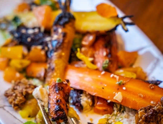 A plate with roasted carrots, pieces of yellow vegetables, herbs, seeds, and a fork. The dish looks colorful and appetizing, set on a wooden table.