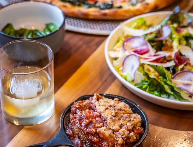 The image shows a tabletop with a pizza, a bowl of fresh salad, a drink with ice, and a small skillet of dessert on a wooden table.