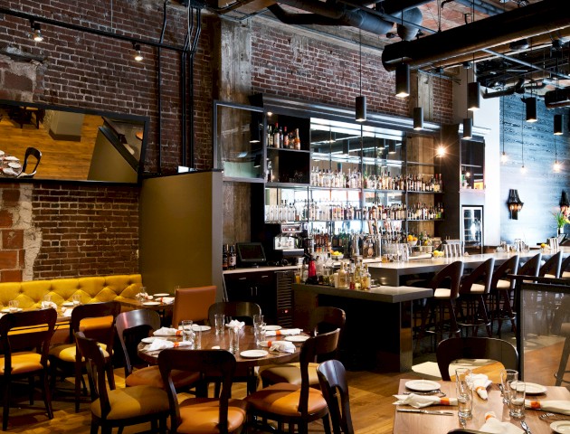 The image shows a modern restaurant with exposed brick walls, a bar, and tables set for dining, creating a warm and inviting atmosphere.