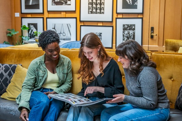 Three people sit on a couch looking at a book, with framed photos on the wall behind them, in a cozy room.