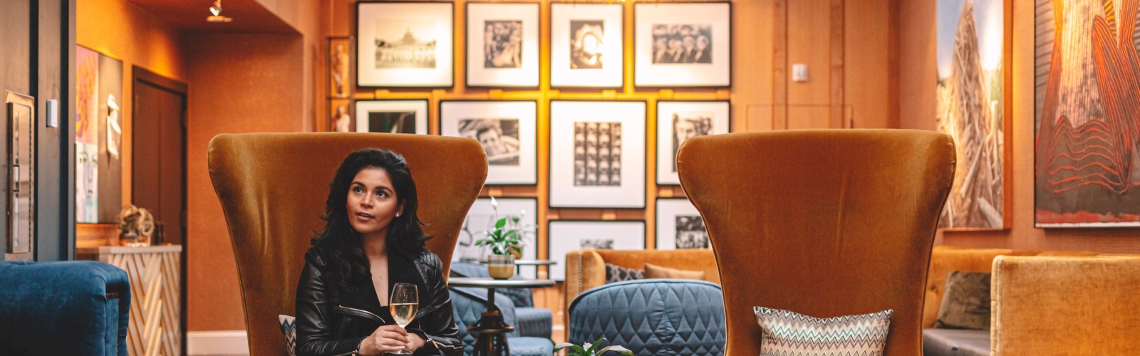 A woman sits in a stylish lounge with high-back chairs, art on the walls, and a warm, inviting ambiance, holding a glass.