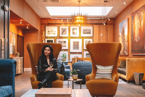 A woman sits in a stylish lounge, holding a drink, surrounded by art and modern furniture in a warmly lit room.