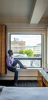 A person sits on a window ledge in a room, gazing out at an urban view. The room has a bed, table lamp, and curtains.