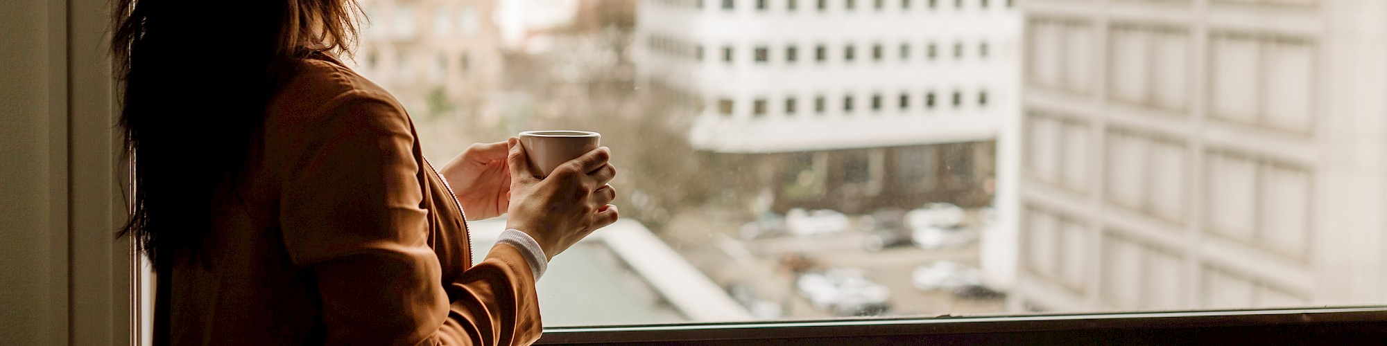 A person stands at a window, holding a mug, gazing out at a cityscape with several buildings in view.