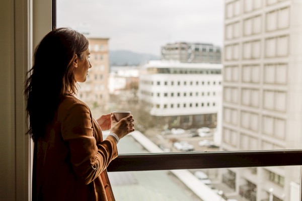 A person holding a cup looks out a window at an urban scene with buildings and a cloudy sky.