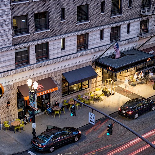 Aerial view of a lively street scene at night with parked cars, outdoor dining tables, and people outside a brick building with glowing lights.