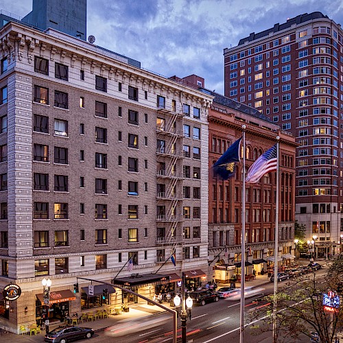 A city street scene with multi-story buildings, illuminated windows, flags, and parked cars under a cloudy evening sky.