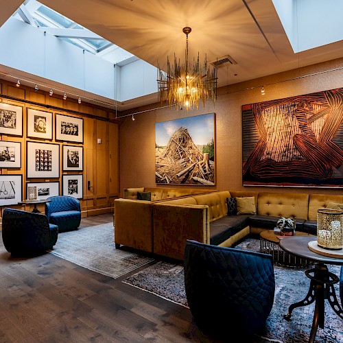 A cozy gallery space features framed art on wood-paneled walls, plush seating, a chandelier, and a skylight for natural light.