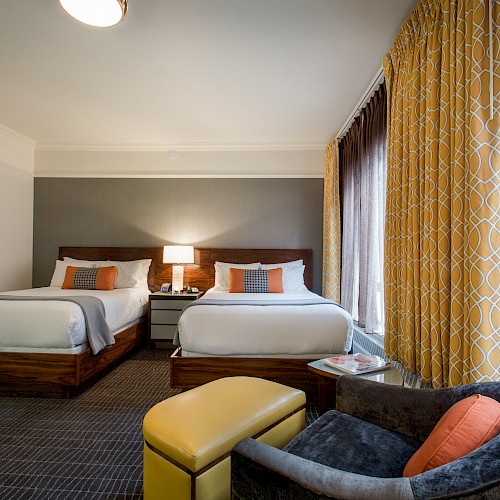 Deluxe Double/Double Room - The image shows a hotel room with two beds, a lamp, yellow and gray decor, a cushioned chair, and a window with patterned curtains.