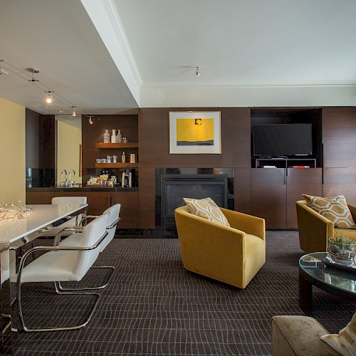 Penthouse Gallery Suite - A modern living room with a dining table, armchairs, artwork on walls, and a TV, featuring neutral tones and a contemporary design.