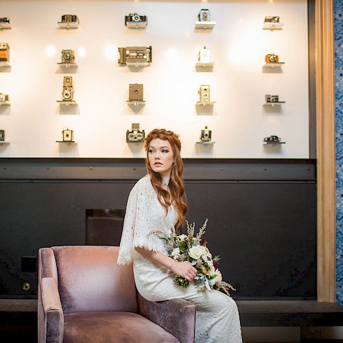 A woman in a white dress sits on a chair, holding a bouquet. Behind her, a wall displays vintage cameras on shelves.