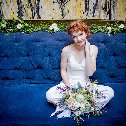 A person with red hair sits on a blue couch, wearing a white outfit and holding a bouquet of flowers, surrounded by greenery and art decor.