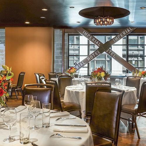 The image shows a restaurant with round tables set for dining, featuring floral centerpieces and a view of the outside through large windows.